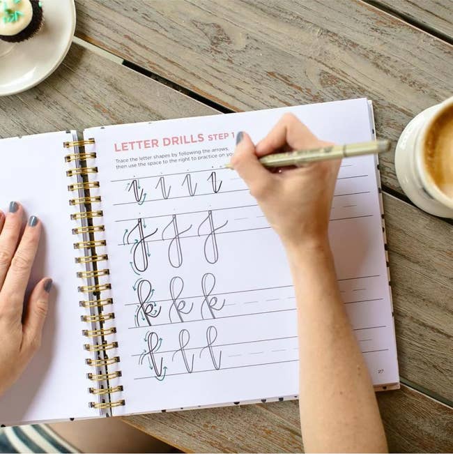 Hand practicing cursive lettering successful  a spiral-bound workbook beside a java  cupful  and cupcake