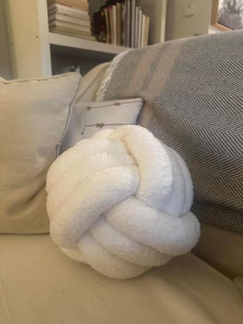 Decorative white knot pillow on a cream couch, with books on a shelf in the background