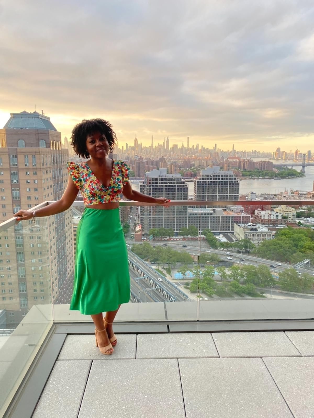 reviewer in a floral top and green skirt standing on a balcony with a city skyline in the background
