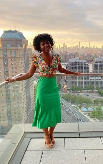 reviewer in a floral top and green skirt standing on a balcony with a city skyline in the background