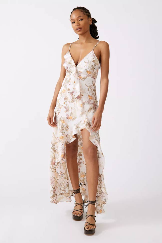 Model wearing white sleeveless dress with purple and orange floral pattern, V-ruffle neckline and a high-low skirt paired with strappy black heels on a white background
