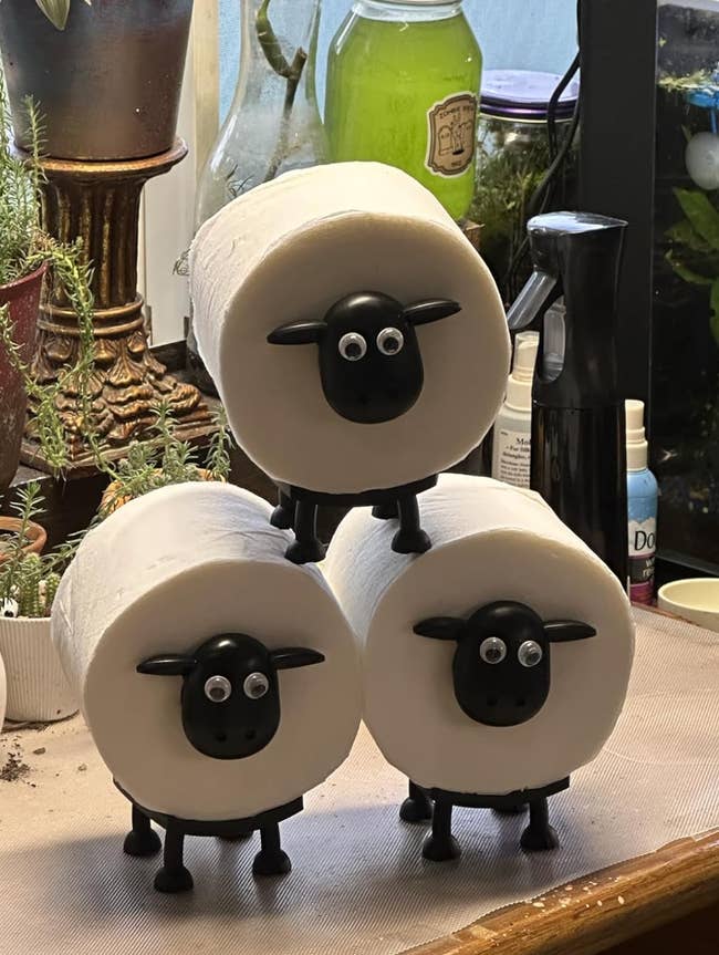 Three toilet paper rolls with playful sheep holders, featuring googly eyes and black faces, stacked on a counter amidst various household items
