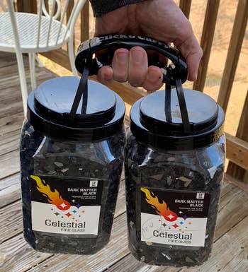 a reviewer photo of a hand holding two ten-pound jars of fire glass using the bag holder 
