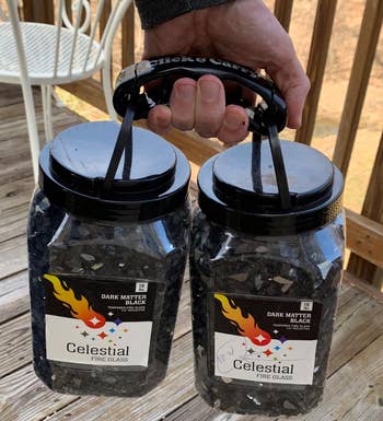 a reviewer photo of a hand holding two ten-pound jars of fire glass using the bag holder 
