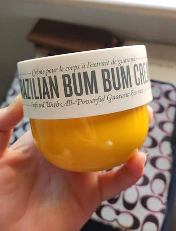 Hand holding a jar of Brazilian Bum Bum Cream with disposable branding and text