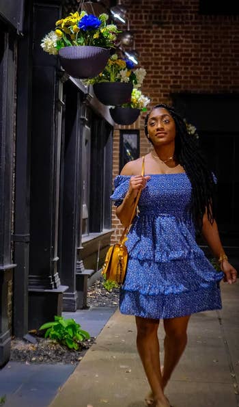 reviewer in a blue ruffled dress with a shoulder bag walking on a city sidewalk at night