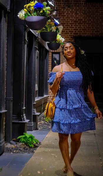 reviewer in a blue ruffled dress with a shoulder bag walking on a city sidewalk at night