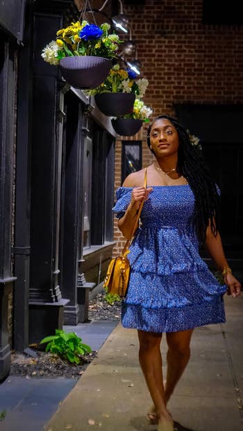 Woman in a blue ruffled dress with a shoulder bag walking on a city sidewalk at night