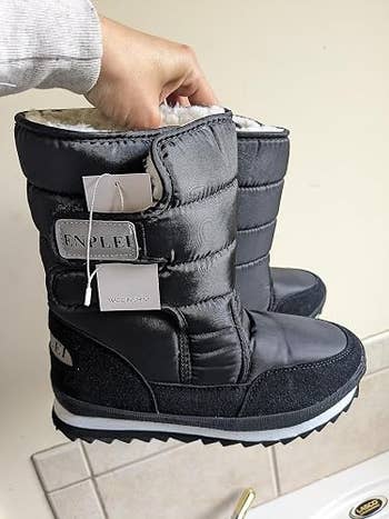 Person holding a brace of insulated wintertime boots with plush lining, featuring tags attached