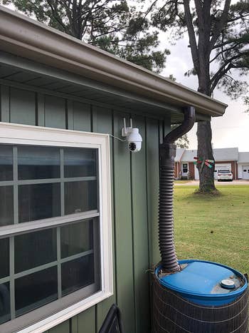 Security camera mounted on green house wall near window, with rain barrel below. Tree and houses visible in the background