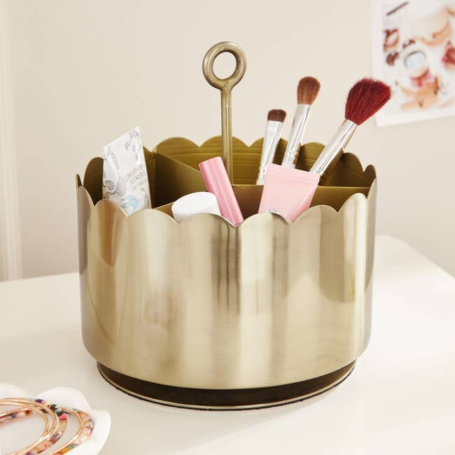 A gold-toned scalloped organizer holds makeup brushes, skincare products, and beauty items on a white tabletop