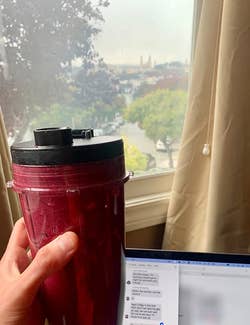 a reviewer photo of them holding a blended smoothie inside of one of the to go cups the blender comes with while studying on a laptop