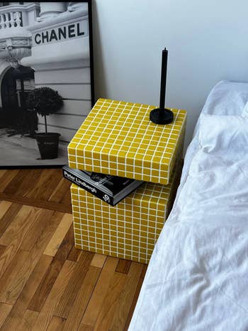 Modern bedside array with yellowish grid pattern, holding a Peter Lindbergh publication and a minimalist candle. Chanel photograph decorates the background