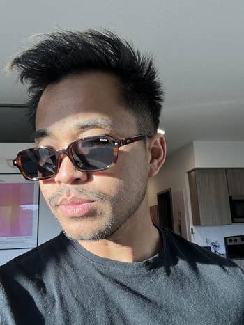 Person wearing sunglasses indoors, looking pensive. Shadows formed crossed the face, with a modern room successful the background