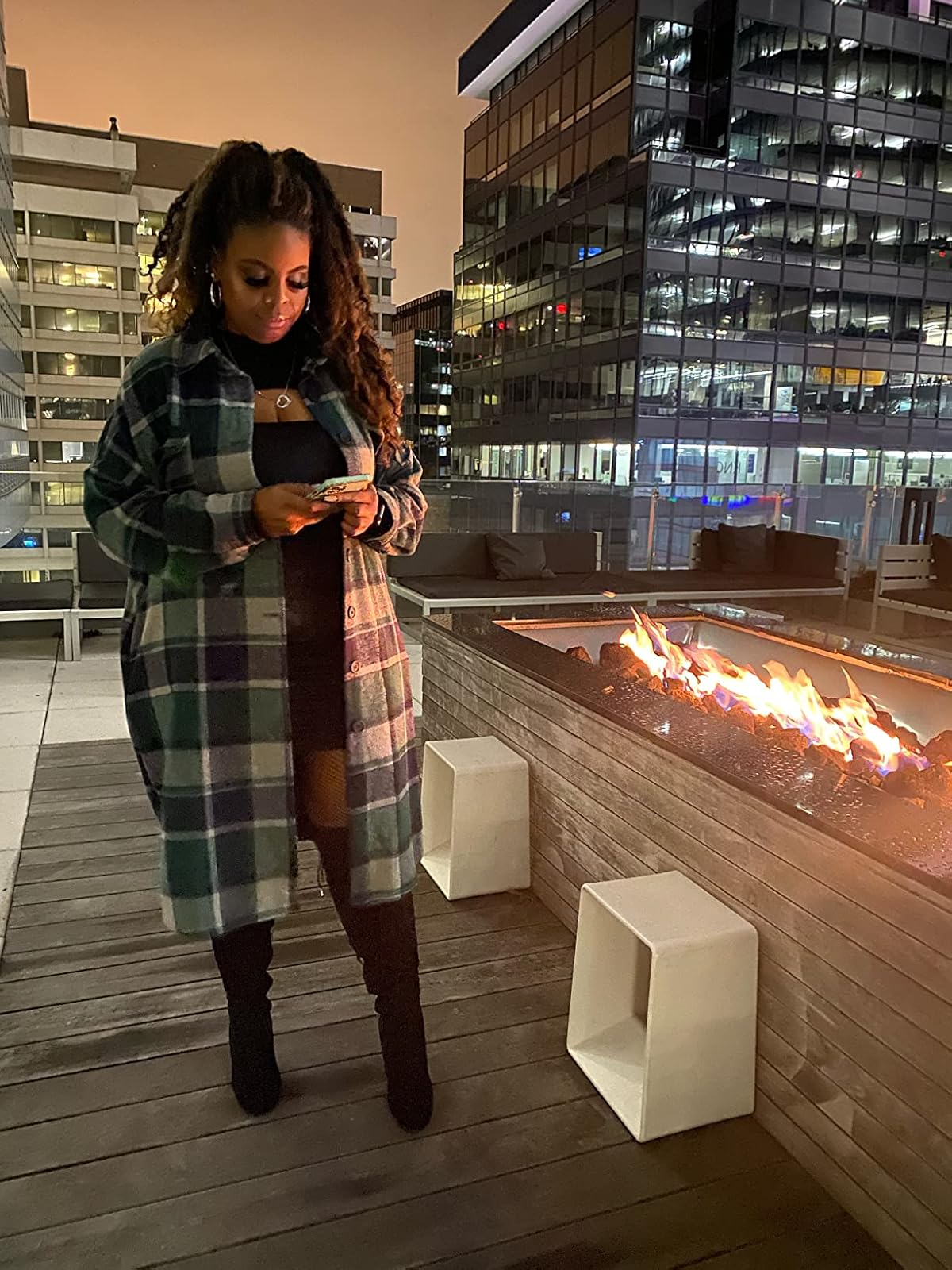 Reviewer successful  a greenish  plaid overgarment  and achromatic  boots stands by a rooftop occurrence  pit, looking astatine  her phone, surrounded by metropolis  buildings astatine  night