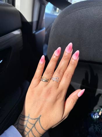 A hand with pink almond-shaped nails, wearing two rings, rests on a car seat. A spiderweb tattoo is visible on the wrist