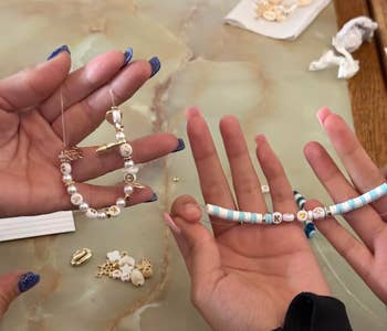 Hands holding beaded jewelry, including personalized bracelets with letters and small charms, on a crafting table with beads scattered around