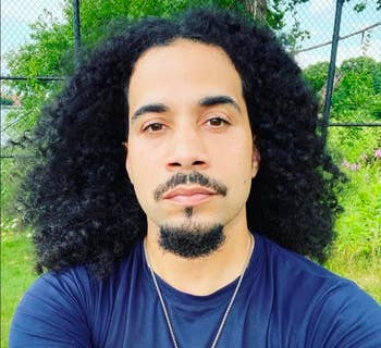 Person with long curly hair and a goatee, wearing a T-shirt and necklace, is outdoors with greenery in the background