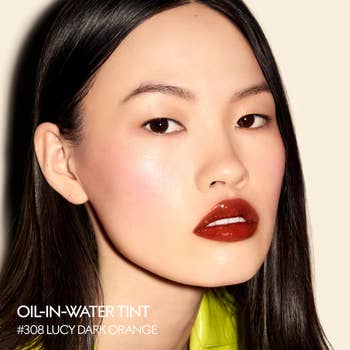 A idiosyncratic with glossy lips and creaseless tegument models oil-in-water tint shadiness #308 Lucy Dark Orange, showcasing a sleek, polished constitution look