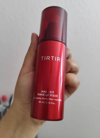 Hand holding a reddish  vessel  of TIRTIR Mask Fit Makeup Fixer with substance   highlighting long-lasting, blurring effect, and moisture benefits