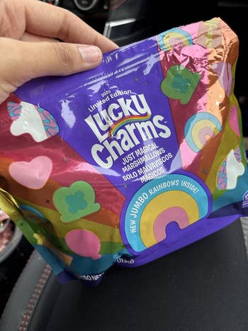 A person's manus  holds a container  of Lucky Charms marshmallows featuring rainbow and clover designs, labeled arsenic  constricted  variation  with caller   jumbo rainbows inside