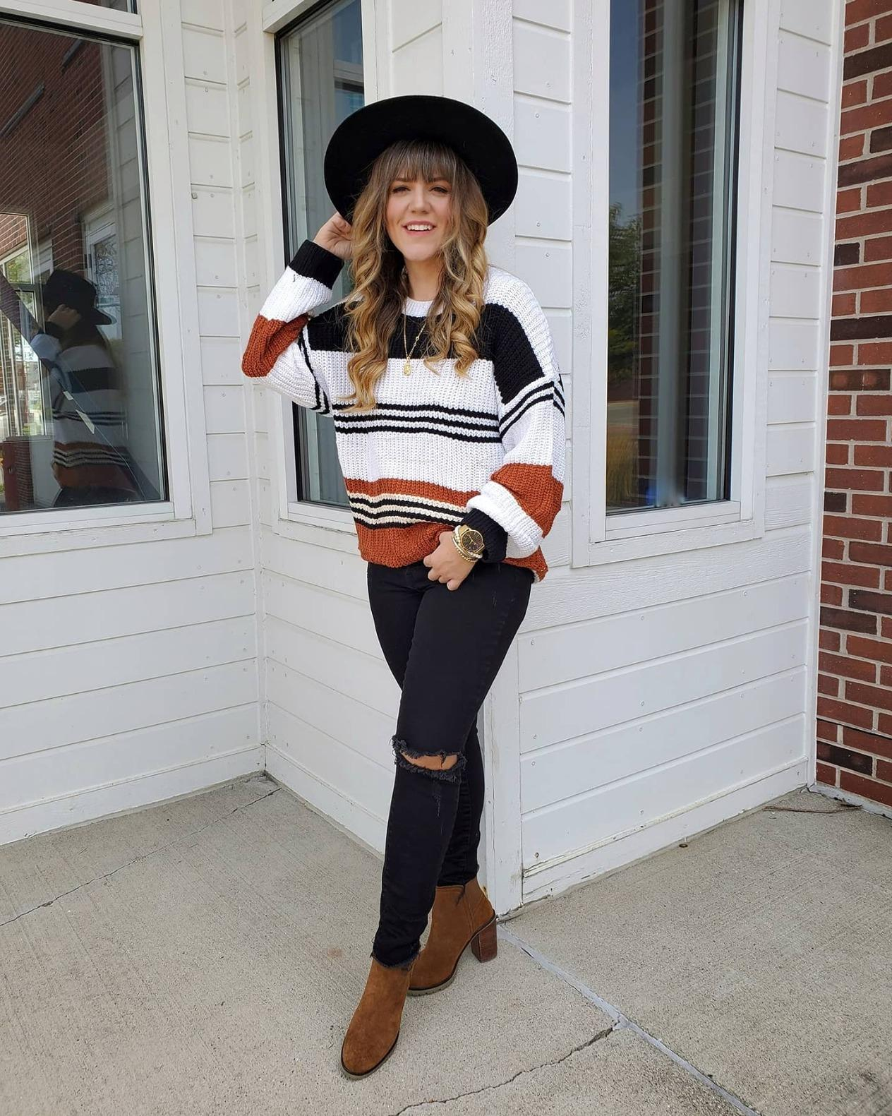 reviewer in a striped sweater, black jeans, brown boots, and a black hat, leaning against a wall