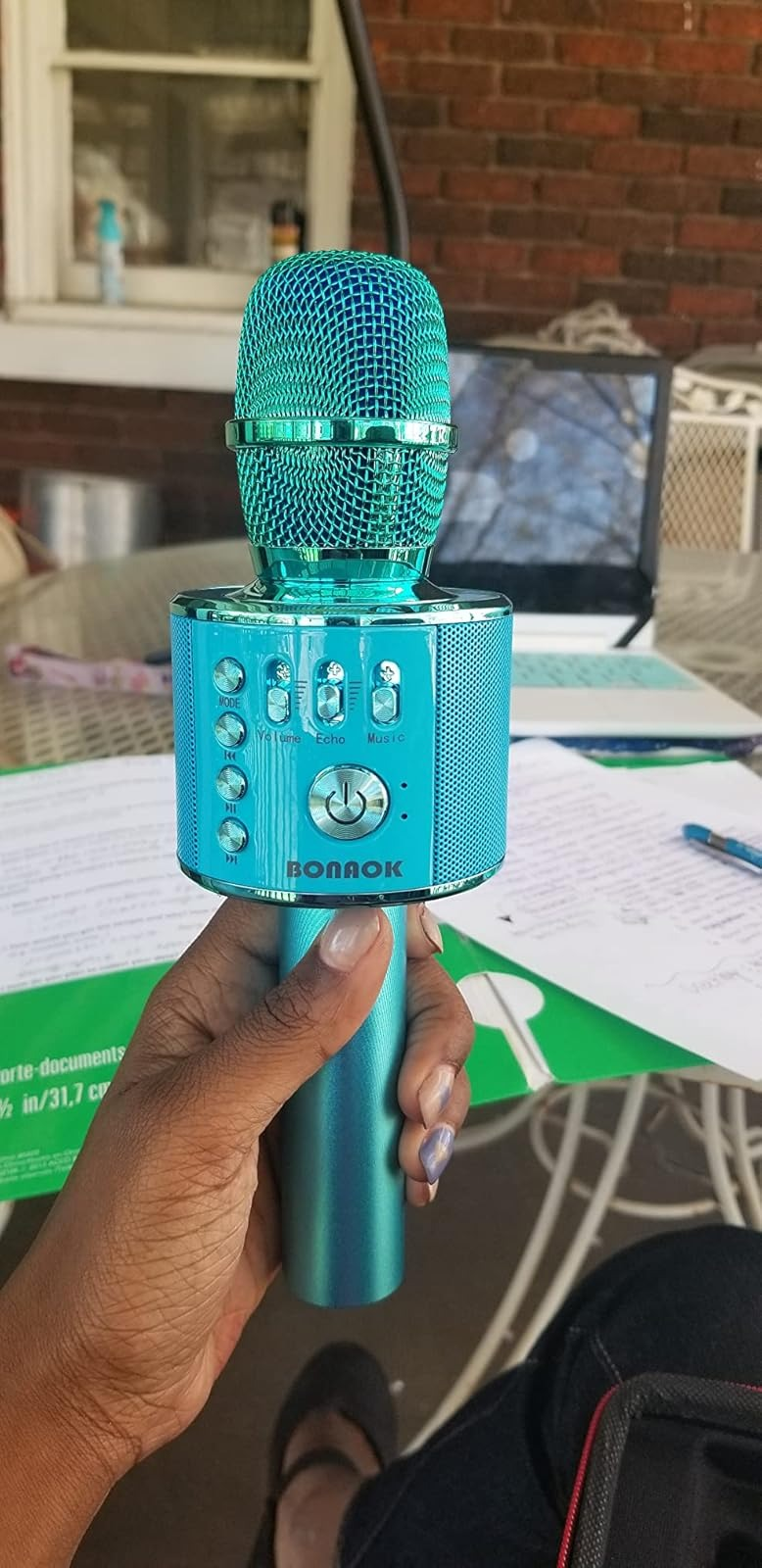 Person holding turquoise karaoke microphone outdoors, with papers and a laptop successful  the inheritance  connected  a patio table