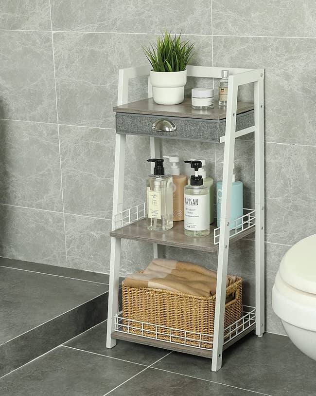 White and brown wooden three-tier ladder shelf with basket and bottles next to the toilet