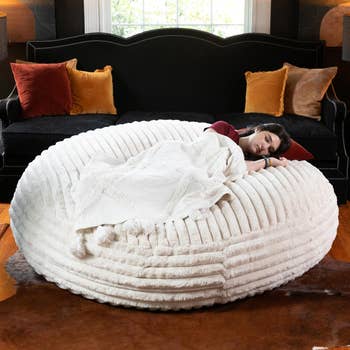 Person sleeping on a large, plush bean bag in a living room with a dark sofa and decorative pillows in the background
