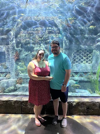Two people stand in front of an aquarium display, featuring fish and coral. The woman wears a dotted dress; the man wears a casual shirt and shorts