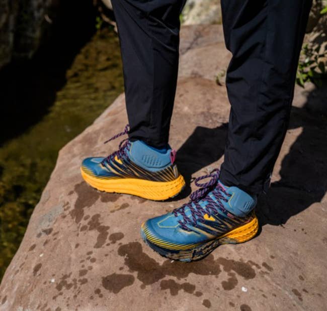 model wearing blue and yellow Hoka hiking shoes on a cliff