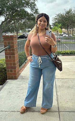 A person takes a mirror selfie, wearing a brown top and wide-leg jeans with a drawstring. They carry a shiny shoulder bag and wear wedge sandals