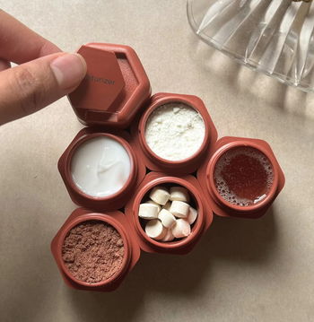 hand placing a labeled lid over one of the red containers, which is stacked with five other containers filled with assorted personal care products