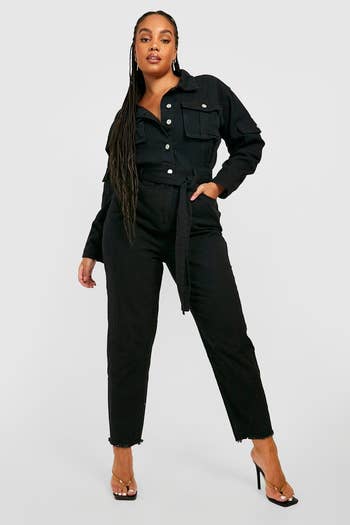 Model posing in a black denim jumpsuit with a cinched waist, paired with black high-heeled sandals