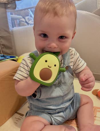 Baby successful  a striped garment  and overalls sitting connected  the floor, teething connected  an avocado-shaped toy