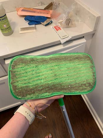 Hand holding a used mop with a green head, showing accumulated dirt, in a household cleaning setting. Various cleaning supplies visible nearby