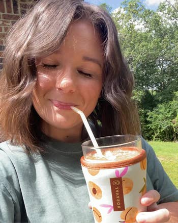 Person enjoying a drink outdoors with a smile, using a reusable straw and holding a glass in a patterned sleeve with fruit designs