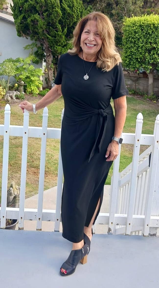 Person outdoors in ankle-length black dress with side tie and peep-toe sandals, standing by a white fence, smiling. Casual chic style