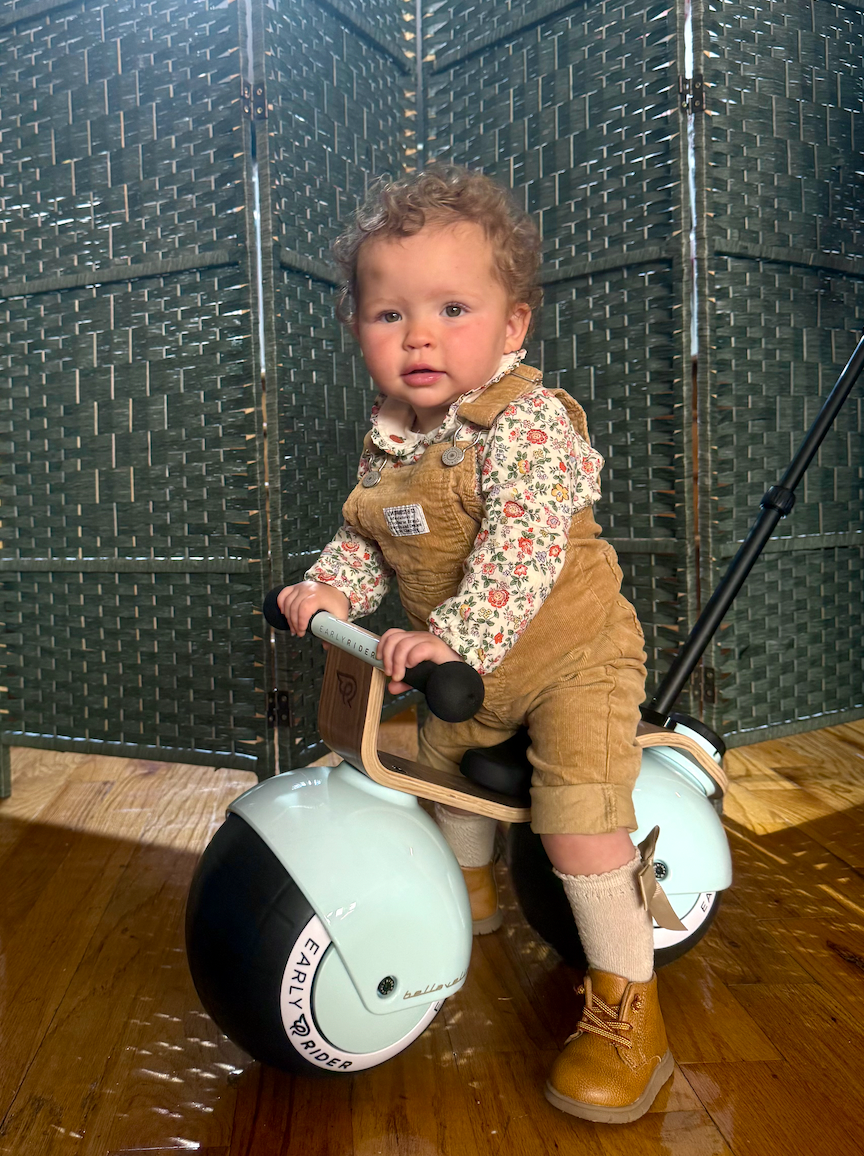Toddler in floral shirt and corduroy overalls sits on a stylish ride-on toy indoors