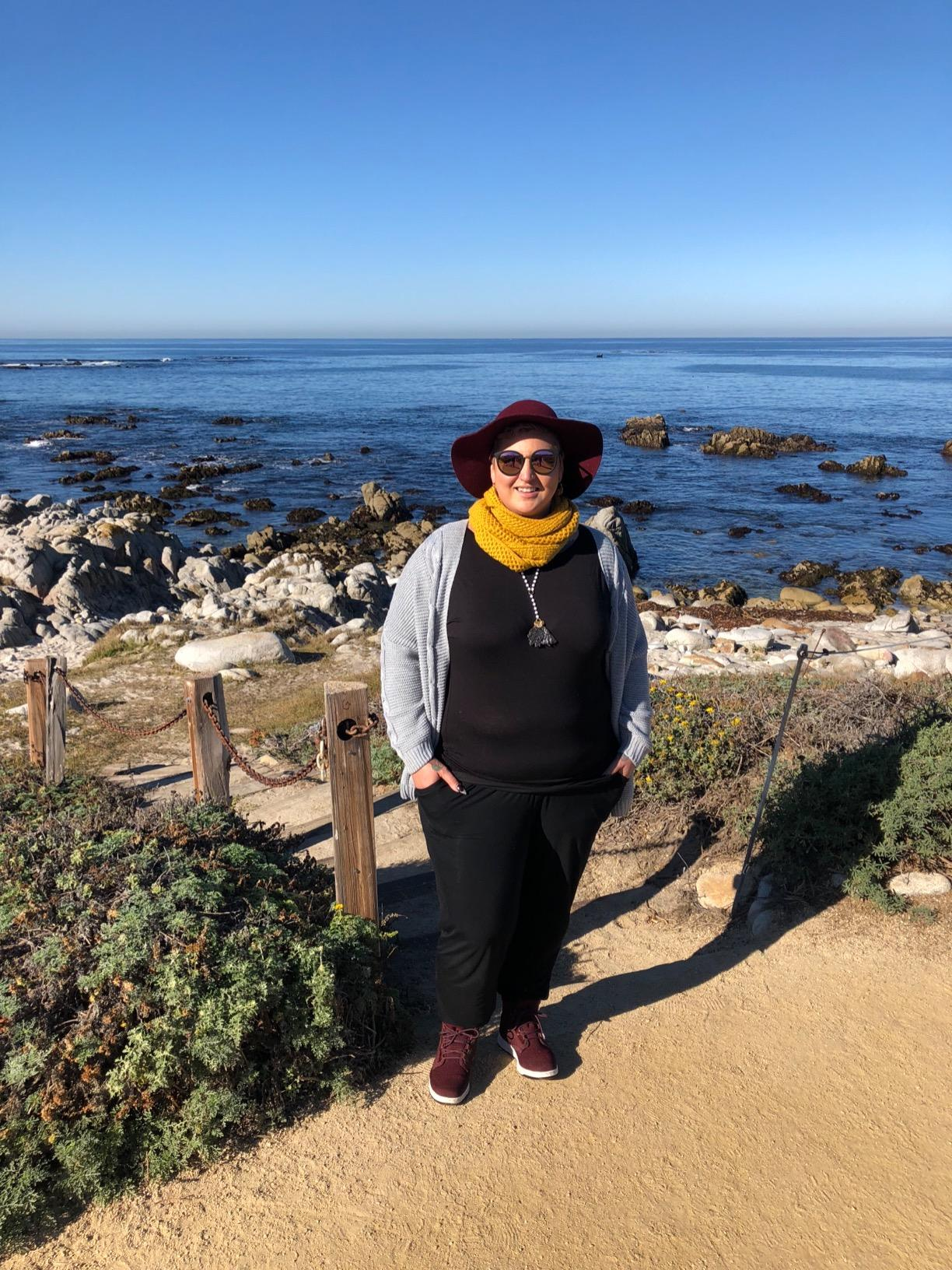 Person smiling by rocky water  view, wearing a wide-brimmed hat, glasses, grey  jacket, scarf, and casual shoes. Perfect for outdoor adventures