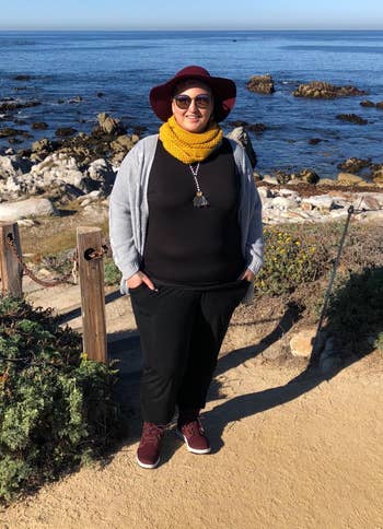Person smiling by rocky water view, wearing a wide-brimmed hat, glasses, grey jacket, scarf, and casual shoes. Perfect for outdoor adventures