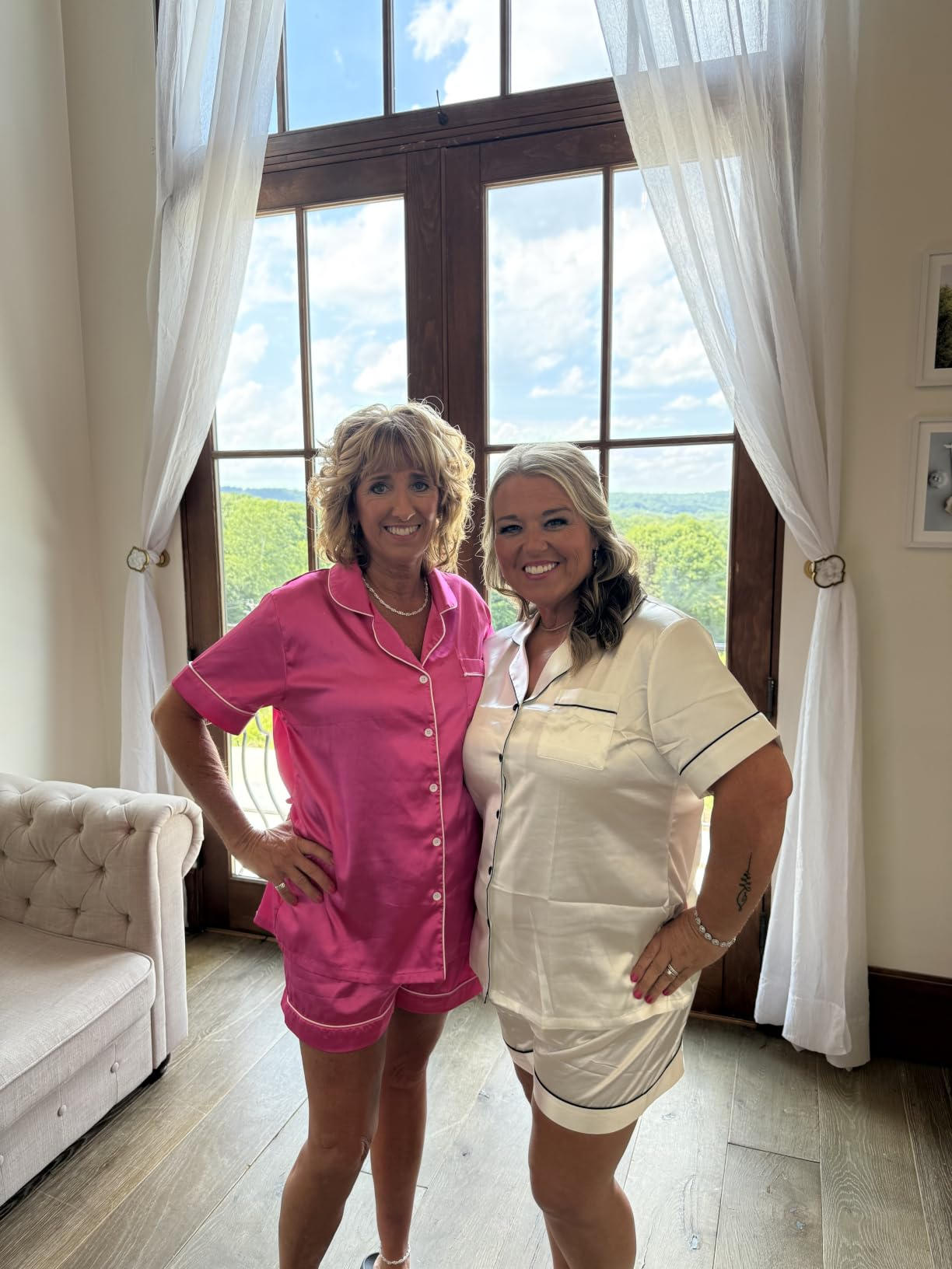 Two women smiling, wearing short-sleeve and shorts pajama sets, basal   indoors by a ample  model   with countryside view