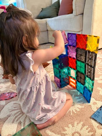 Toddler playing with colorful magnetic gathering  blocks connected  a patterned rug adjacent   a sofa