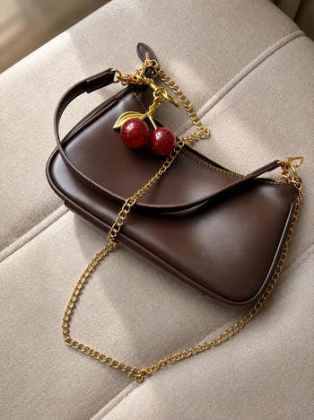 Brown handbag with a gold chain strap and cherry charm, placed on a beige fabric surface