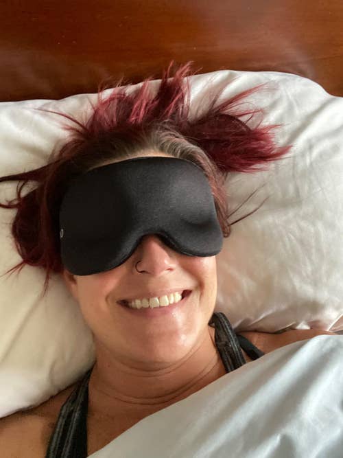 a reviewer wearing the black sleep mask in bed while smiling 
