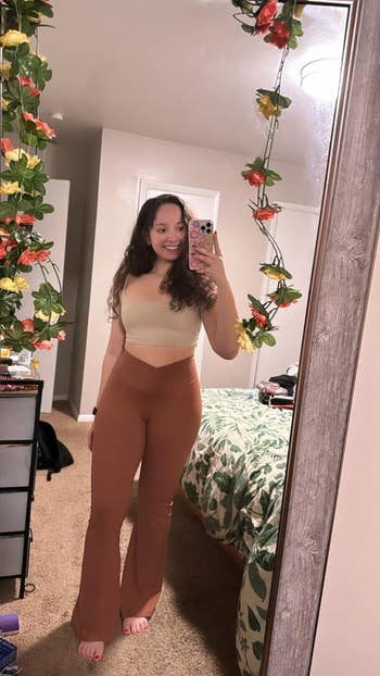 Orang yang mengambil selfie cermin di kamar tidur, mengenakan atasan atas dan celana berpinggang tinggi, dengan dekorasi karangan bunga yang terbungkus di sekitar cermin