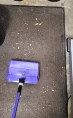 a purple flat broom with bristles picking up crumbs from a carpet 