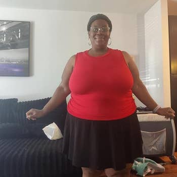 Person smiling indoors, wearing a sleeveless apical and skirt, lasting successful a surviving country with a sofa and partition art