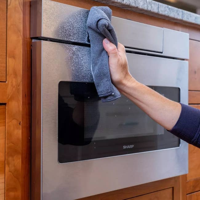 hand wiping a stainless-steel oven with the cleaner using a microfiber cloth