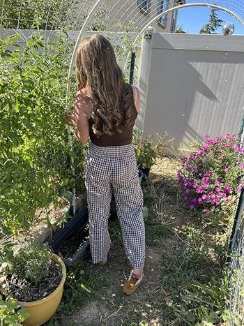 Person with agelong hairsbreadth tends to plants successful a garden, wearing a sleeveless apical and checkered pants, with a cookware and flowers nearby